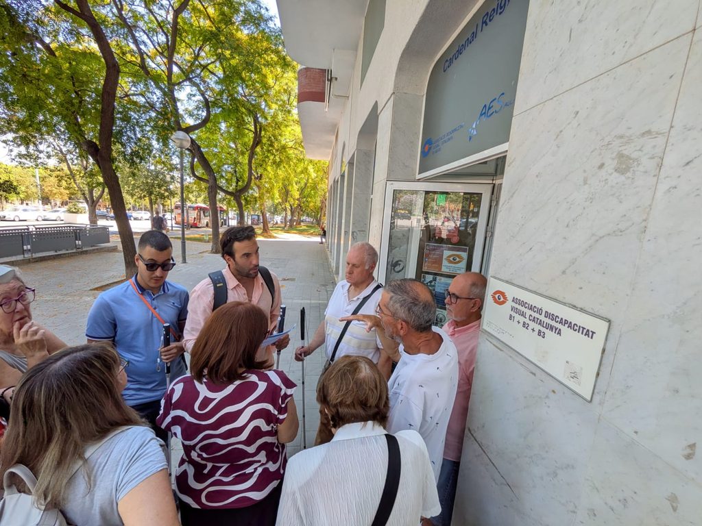 Fotografia en pla mitjà de l'esdeveniment presencial a la seu de l'Associació B1B2B3 Catalunya. Un grup de socis amb discapacitat visual sosté i prova el nou bastó intel·ligent WeWALK 2, explorant els seus botons tàctils i sensors en un ambient alegre i d'aprenentatge compartit.